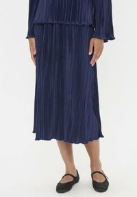Jupe plissée navy au design fluide, dotée d'un ourlet en dentelle, associée à des ballerines noires. Le tissu a un aspect doux et texturé.