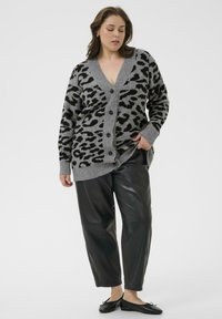Grauer Cardigan mit Leopardenmuster, V-Ausschnitt und Knopfverschluss; hat eine dicke Stricktextur. Kombiniert mit schwarzen Lederhosen und flachen Schuhen.