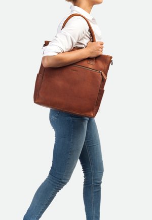 Bruine leren tote bag met dubbele handgrepen, gestructureerd oppervlak, afsluitbare bovenkant met rits en zijzakken, gedragen op de schouder tegen een lichte achtergrond.