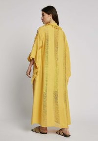 Losse gele kaftan met franjes en verticale strepen. Zachte stof, mouwen van gemiddelde lengte en zijopeningen voor comfortabele bewegingsvrijheid.