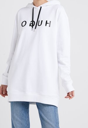 Osoba ubrana w biały, oversize'owy kaptur z czarnymi sznurkami i odwróconymi czarnymi literami na wysokości klatki piersiowej, w połączeniu z niebieskimi dżinsami.