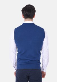 Maglione senza maniche blu da uomo realizzato in tessuto lavorato a maglia, con scollatura a girocollo e orlo a costine. Indossato sopra una camicia bianca.