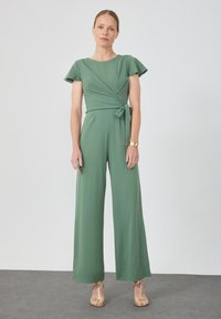Grön jumpsuit med korta volangärmar, omslagningsdesign, knytning i midjan och vid ben. Slät tyg med matt yta.