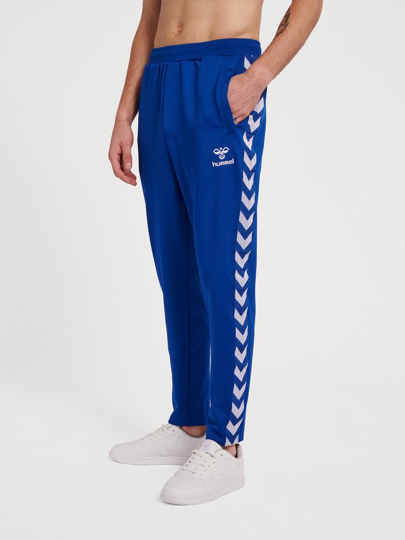 Homme portant un pantalon de survêtement bleu Hummel avec des bandes chevrons blanches sur les côtés, assorti à des baskets blanches, debout avec une main dans la poche.