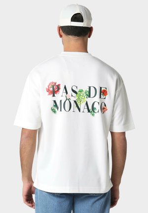 Hvid t-shirt med korte ærmer, som har blomstergrafik og teksten "PAS DE MONACO" i sort. Stoffet er glat, og skjorten har en afslappet pasform.