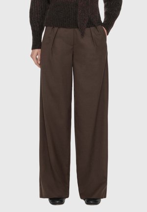 Brown wide-leg trousers with front pleats worn with a dark knitted sweater and black shoes, hands in pockets.