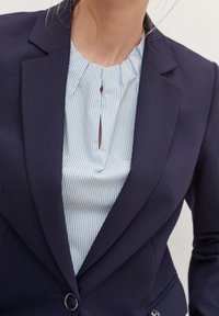 Granatowy żakiet z wyraźnymi klapami na lekko niebieskiej bluzce w paski białe z małym otworem w kształcie klucza przy dekolcie.