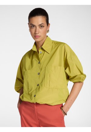 Frau mit zurückgekämmtem Haar, trägt ein locker sitzendes, limettengrünes Hemd mit Knöpfen und korallenfarbene Hosen, Hände in den Taschen, vor weißem Hintergrund.