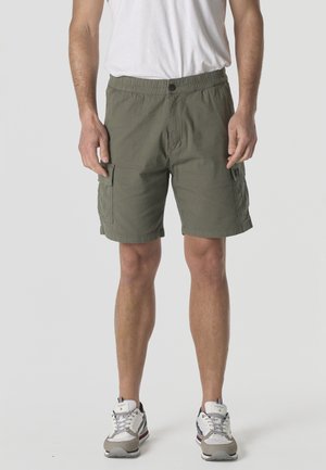 Hombre con pantalones cortos cargo verde oliva con bolsillos laterales, camiseta blanca y zapatillas blancas, de pie contra un fondo liso.