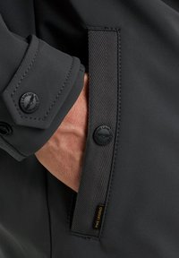 Black fabric jacket detail showing a hand in the pocket, featuring a textured accent band, button closure, and a logo patch.