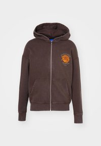 Brun zip-up hoodie med en känguruficka, dragsko i huven och en orange grafisk design på vänster bröst. Mjuk tygtextur.