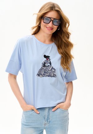 Kobieta o długich falowanych włosach, nosząca duże okulary przeciwsłoneczne, jasnoniebieską koszulę z abstrakcyjnym wzorem oraz jasnoniebieskie dżinsy, ręce w kieszeniach.