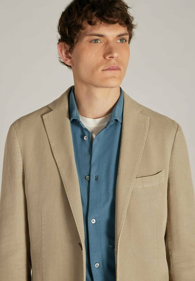 Beige linnen blazer met een notch lapel en een enkele borstzak, gedragen over een blauw shirt met knopen en een ronde hals.