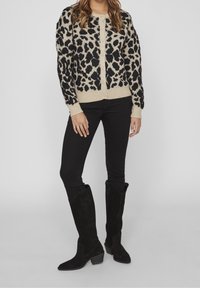 Cardigan imprimé léopard beige et noir, avec fermeture à boutons, poignets et corps côtelés. Associé à un jean noir ajusté et des bottes hautes noires.
