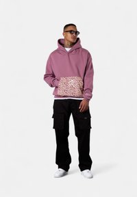 Pink pullover hoodie with leopard print pocket, paired with black cargo pants. White sneakers complete the look. Smooth texture, relaxed fit.