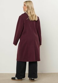 Burgundy takki pitkillä hihoilla ja suoralinjaisella leikkauksella, jossa on sileä pinta. Yhdistetty mustiin leveälahkeisiin housuihin ja urheilullisiin mustiin kenkiin.