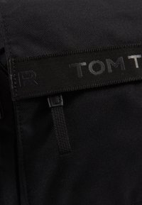 Zaino nero realizzato in un tessuto resistente, con una nastro logo prominente e una superficie testurizzata caratterizzata da elementi di design minimalisti.