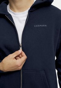 Mörkblå zip-up hoodie i mjukt material, med en silverdragkedja, framfickor och en vit logotyp "CASPARA" på vänster bröst.
