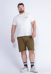 Bílý trikoton s kulatým výstřihem a malým modrým potiskem, kombinovaný s olivově zelenými šortkami. Model má na sobě bílé tenisky a námořnické pruhované ponožky.