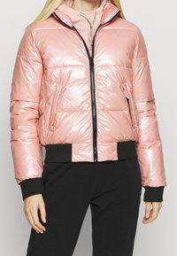 Rosa puffjacka med en glänsande, vadderad yta, frontdragkedja och svarta ribbade muddar vid nederkant och ärmar. Har sidofickor.