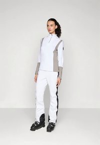 Vrouw in een wit en grijs skikostuum met zwarte skischoenen, staande tegen een effen lichte achtergrond.