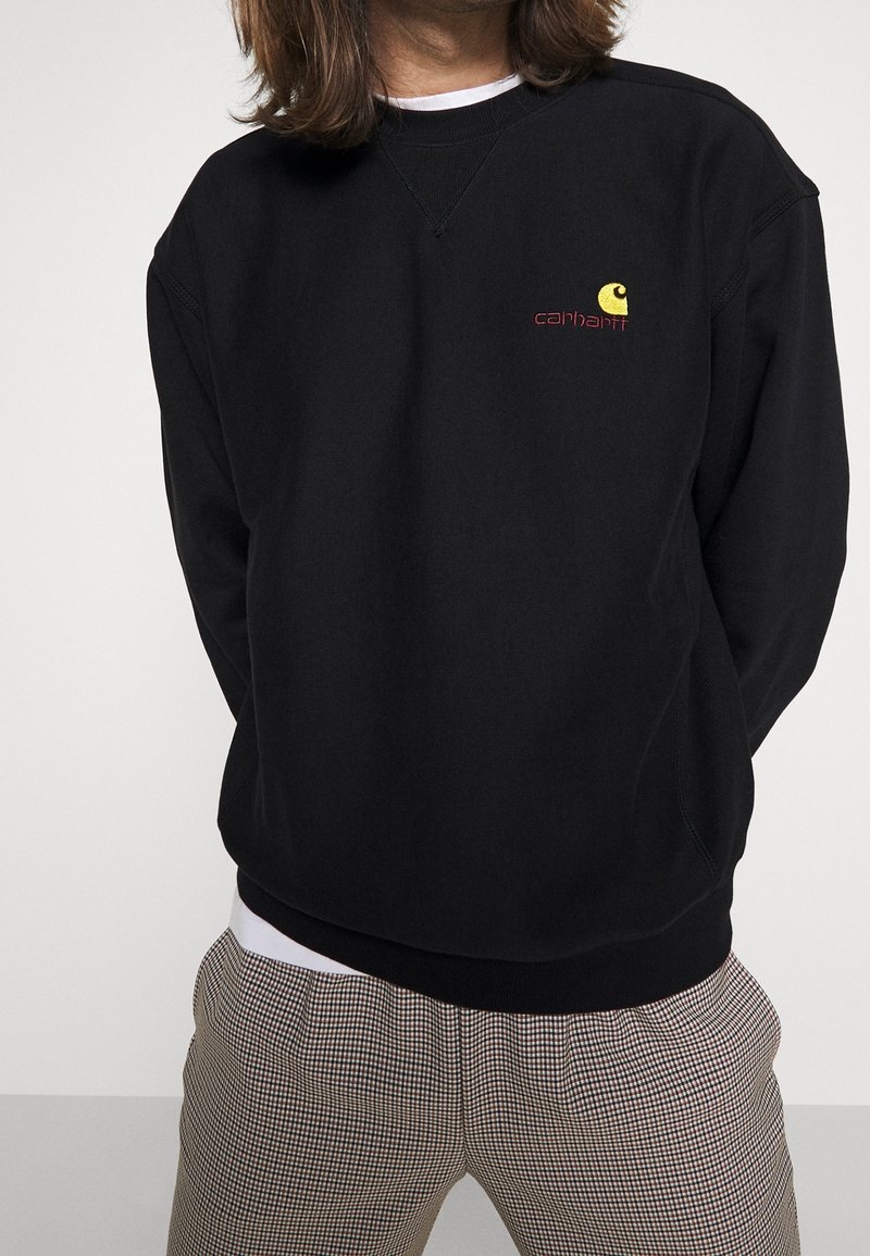 Schwarzes Sweatshirt mit Rundhalsausschnitt und geripptem Saum, versehen mit einem kleinen gelb-roten gestickten Logo auf der Brust. Getragen über karierten Hosen.