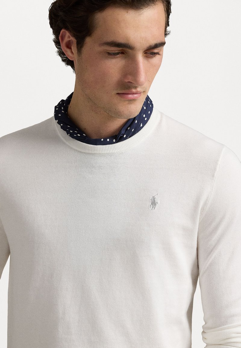 Suéter de punto blanco con cuello redondo, que presenta un pequeño logo bordado en plata en el pecho. Combinado con una bufanda de lunares azul marino.
