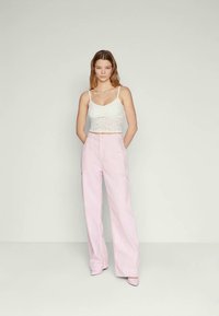 Weißes Spitzen-Crop-Top und hellrosa weit geschnittene Hose mit vorderen Taschen und glatter Textur. Modell steht vor einem neutralen Hintergrund.