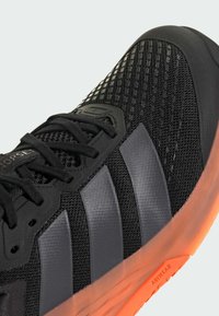 Svart sportssko med mesh-overdel, syntetiske overlapp og tre grå striper. Har en gjennomsiktig oransje gummisåle.