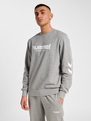 Mand iført grå Hummel sweatshirt og matchende joggingbukser med hvidt logo og chevron-design, stående mod ensfarvet baggrund.