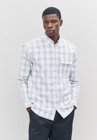 Camicia a quadri azzurro chiaro e bianca con bottoni, realizzata in tessuto dalla texture morbida. Presenta una tasca sul petto e un colletto standard.