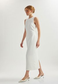 Abito midi bianco con una linea elegante, caratterizzato da un collo alto, spacchi laterali e una texture liscia. Abbinato a sandali con tacco bianchi.