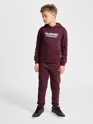 Chłopiec z kręconymi włosami stoi i patrzy na lewo, ubrany w bordową bluzę z kapturem hummel, pasujące spodnie dresowe i czarne trampki na białym tle.