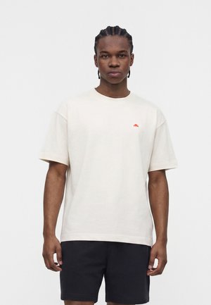 Noor mees punutud juustega, kandes tavalist valget t-särki väikese oranži logoga ja musti lühikesi pükse, lihtsa taustaga.