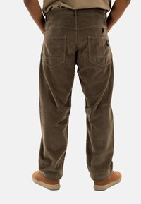 Pantalones de pana marrones con una textura acanalada, que presentan bolsillos traseros y un ajuste relajado. Combinados con zapatos marrones sobre un fondo claro.