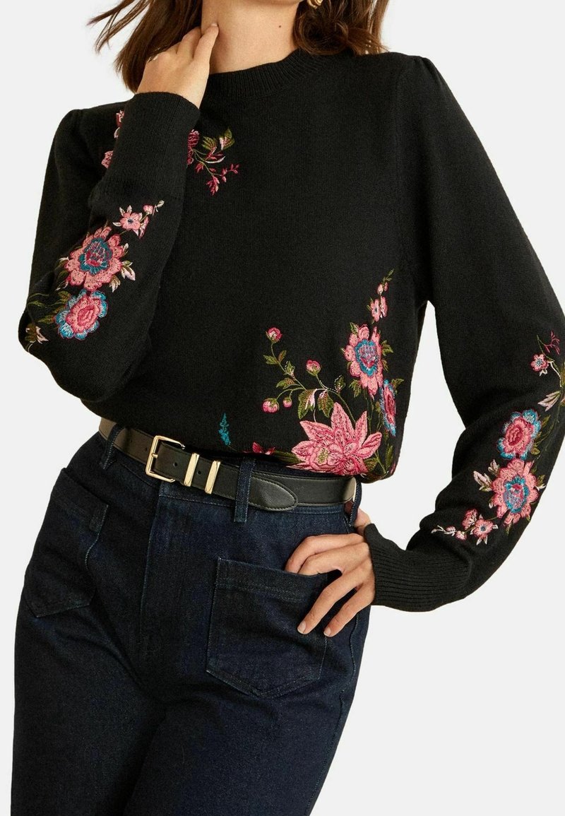 Femme portant un pull noir avec une broderie florale rose et bleue sur les manches et le devant, associé à un jean foncé et une ceinture noire.