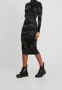 Klänning med kamouflagemönster, lång ärm i mörkgrön och svart. Slim fit, längd till mitten av vadderna, kombinerad med svarta snörkängor och gula detaljer.