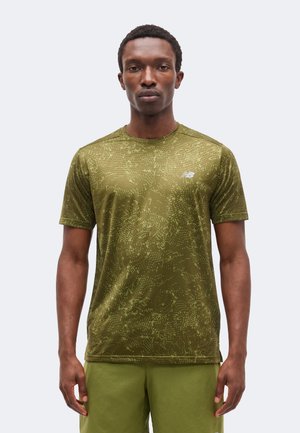 Hombre con camiseta deportiva de manga corta de color verde oliva con estampado y pantalones cortos a juego, de pie frente a un fondo liso y claro.