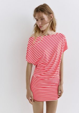 Junge Frau mit langen Haaren, die ein kurzes rot-weiß diagonal gestreiftes Kleid trägt, stehend vor einem weißen einfarbigen Hintergrund.