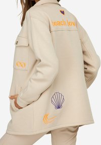 Veste en polaire beige au design décontracté, brodée du texte "beach lov" en orange, avec un cœur violet, un symbole d'infini, une coquille et des détails de vague.