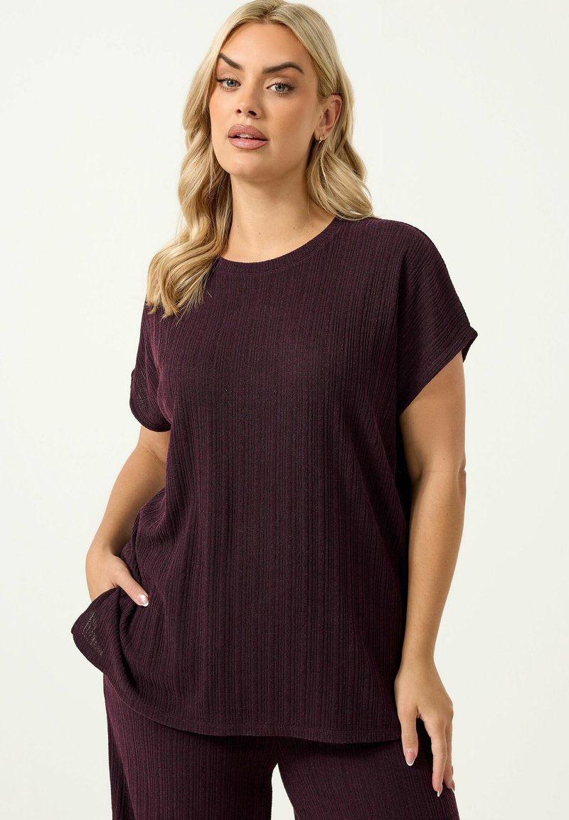 Donna con capelli biondi che indossa una maglia corta a maniche corte larga a coste di colore viola scuro e pantaloni abbinati, in piedi con una mano in tasca.