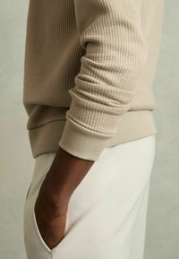 Beige ribbet sweater med en tætsiddende manchet, parret med lyse bukser. Nærbillede af den teksturerede ærme og lommeomrids.