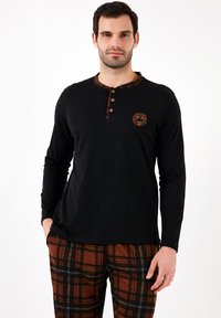 Camisa negra de manga larga con cuello de cuadros, con tres botones y un logo redondo. Combinada con pantalones de cuadros marrones con acentos blancos.
