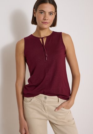 Femme aux cheveux courts et bruns porte un haut bordeaux sans manches avec un détail noué à l’avant et un pantalon beige, posant avec une main dans la poche.