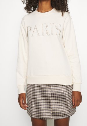 Mujer vistiendo una sudadera crema con texto "PARIS" y una minifalda pata de gallo en blanco y negro, de pie con los brazos relajados.