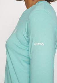 Mintgrünes Langarmshirt aus glattem Stoff. Verfügt über ein weißes Logo auf dem linken Arm und ein kleines Emblem in der Nähe des Halsausschnitts.