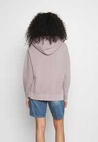 Světle fialová mikina s kapucí na šňůrku, žebrovanými manžetami a spodním lemem. Na sobě s džínovými šortkami, ukazující pohodlný a uvolněný střih zezadu.