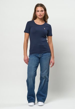 Ung kvinde stående iført en navyblå tætsiddende T-shirt med korte ærmer, blå udvidede jeans og hvide sneakers mod en ensartet lys baggrund.