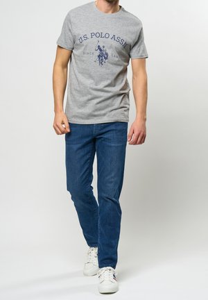 Homme portant un t-shirt gris U.S. Polo Assn., un jean bleu et des baskets blanches, marchant devant un fond clair uni.