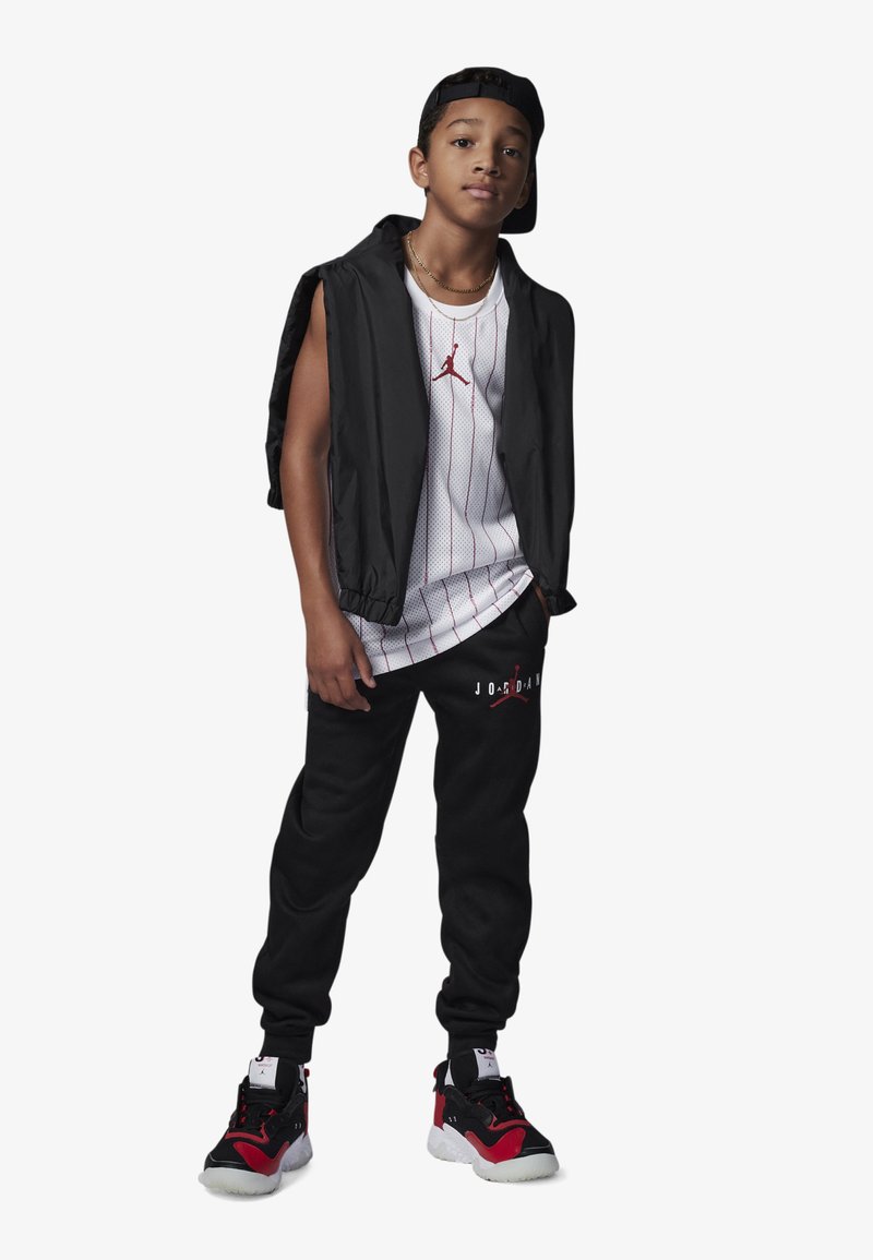 Veste sans manches noire sur un débardeur blanc à rayures avec un logo rouge, joggers noirs avec du texte blanc, et des baskets noires et rouges.